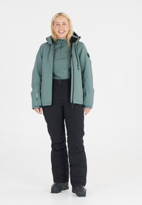 Lichtgroene waterdichte jas over een donkere top, gecombineerd met zwarte geïsoleerde broek en zwarte skischoenen, met een ontspannen pasvorm en ritsdetails.