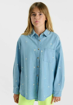 Jonge persoon met lang blond haar, gekleed in een loszittend lichtblauw overhemd met kleine witte hartjespatronen.