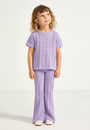 Barn med krøllet hår iført et matchende lavendel farvet strikket sæt bestående af top og bukser, står op og kigger let opad mod højre.
