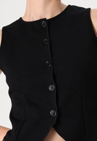 Μαύρη αμάνικη μπλούζα με στρογγυλή λαιμόκοψη, διαθέτει κουμπιά στο μπροστινό μέρος, δομημένο σχέδιο και διακριτή υφή. Χωρίς ορατά σχέδια ή διακοσμήσεις.