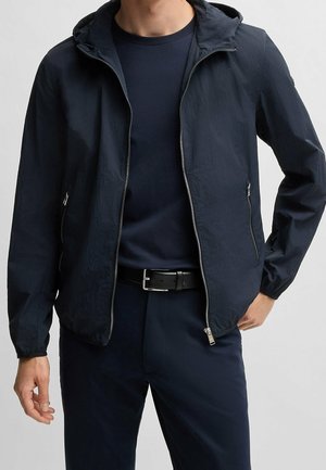 Mann, der eine marineblaue Kapuzenjacke mit Reißverschluss über einem passenden marineblauen Shirt und Hose mit schwarzem Gürtel trägt, steht mit einer Hand in der Hüfte.