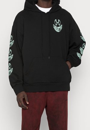 Person iført sort Carhartt WIP hoodie med mintgrønt logo på bryst og ærmer, kombineret med mørkerøde bukser.
