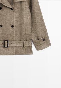 Beige uld dobbeltradet frakke med sorte knapper, bælte i taljen, sidelomme og knaplukket manchet på hvid baggrund.