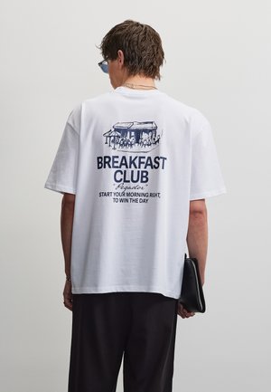Személy fehér pólót visel kék kávézói illusztrációval és a „Breakfast Club, Start your morning right, to win the day“ szöveggel, fekete kézitáskát tartva.