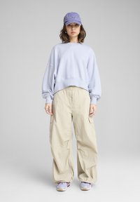 Sweatshirt surdimensionné bleu clair, pantalon cargo beige avec poches latérales, et casquette violette. Les chaussures sont de style athlétique violet et blanc.