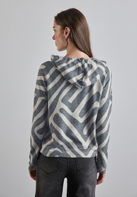 Huvtröja med ett grått och krämfärgat abstrakt mönster, stickad textur och långa ärmar, kombinerad med mörkgrå jeans.