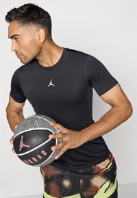 Sort kortærmet atletisk skjorte med et hvidt Jordan-logo, parret med farverige mønstrede shorts og holder en struktureret basketball.