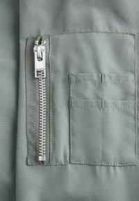 Tessuto verde oliva con una superficie liscia, dotato di una zip argentata a sinistra e di una piccola tasca rettangolare sopra di essa.