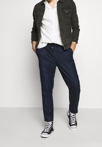 Tmavě šedá džínsová bunda přes bílou košili s krátkým rukávem, doplněná o navy volné kalhoty a černé kotníkové tenisky.