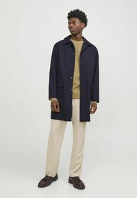 Námornícky trench coat s gombíkovým zapínaním, dvoma prednými vreckami, skombinovaný so béžovým sveterom a smotanovými nohavicami, doplnený tmavými topánkami.