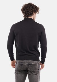 Maglione nero realizzato in morbido tessuto a maglia con un design aderente, maniche lunghe e polsini a costine. Indossato sopra jeans grigi in denim.