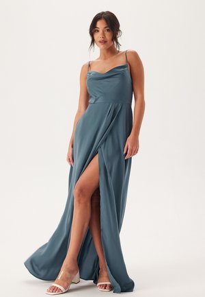 Kappa-halsad, teal satin maxiklänning med en sidoslits. Har spaghettistövlar och en flödande design. Kombineras med neutrala blockklacksandaler.