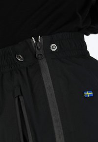 Mustat vedenpitävät housut, joissa on vetoketjullinen etuosa, joustava vyötärö ja pieni ruotsalainen lipputunnus lähellä taskua.