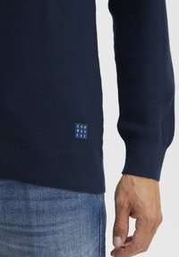 Mornarsko modra pulover z teksturiranim pletenjem, rebrastimi manšetami in robom, z majhno blagovno znamko na strani. Obut z džins hlačami.