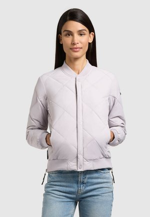 Frau mit langen dunklen Haaren, die eine helllavendelfarbene gesteppte Jacke und blaue Jeans trägt und mit den Händen in den Jackentaschen steht.