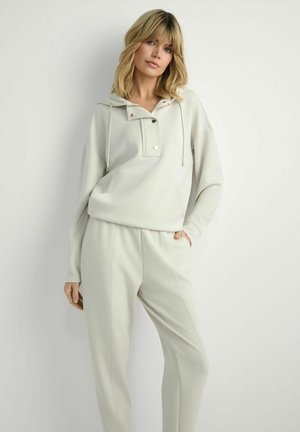 Blondynka w jasno beżowej bluzie z kapturem z guzikami i dopasowanych, luźnych spodniach, stojąca na tle białej ściany.