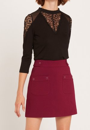 Femme portant un haut en dentelle noire avec des manches trois-quarts et un col montant, associé à une jupe rouge foncé taille haute avec poches avant.
