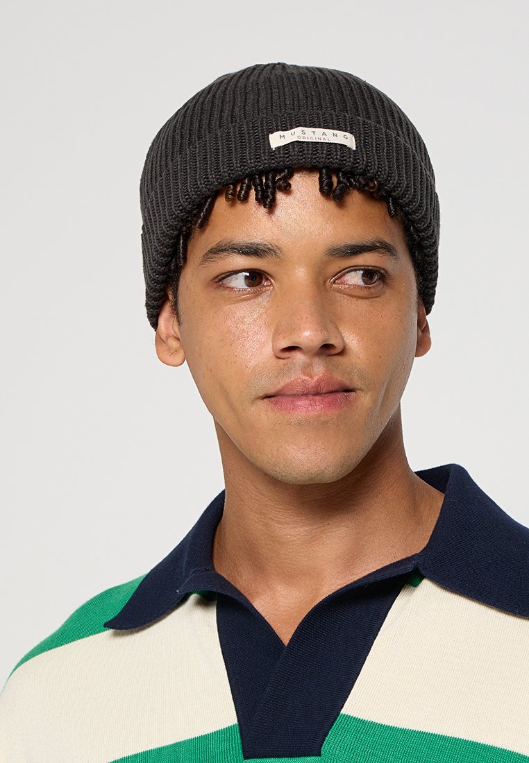Czarna, dziana czapka z wywiniętym mankietem, z kremową metką z przodu. Nosi ją model z krótkimi, ciemnymi lokami.