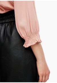 Chemise à manches bouffantes en tissu rose pâle avec un motif en chevrons, dotée de poches élastiquées à volants, associée à une jupe en cuir noir.