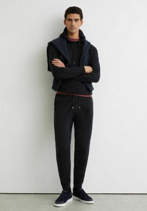 Junger Mann steht mit verschränkten Armen, trägt schwarzen Kapuzenpullover, passende Jogginghosen, marineblaue Sneakers und einen dunklen Pullover über den Schultern, vor einer weißen Wand.