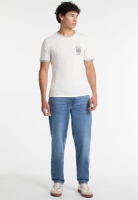 Camiseta de algodón color crema con ribete gris, que presenta un pequeño logo azul en el pecho. Combinada con jeans de mezclilla azul claro y zapatillas beige.