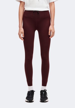 Personne portant un legging moulant bordeaux foncé avec des poches latérales, un t-shirt blanc, des chaussettes blanches et des baskets noires, debout devant un fond clair.