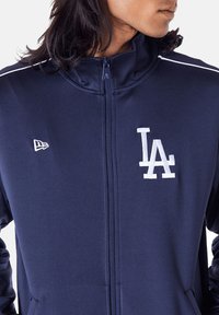 Marineblaue Zip-Up-Jacke mit weißem, gesticktem "LA"-Logo, leichtem Stoff, hohem Kragen und feinem Nähen entlang der Schulternähte.
