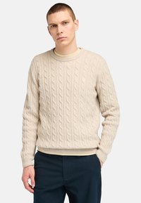 Beige Kabelstrickpullover mit rundem Ausschnitt, gerippten Manschetten und Saum. Über einem hellen Unterhemd getragen, kombiniert mit dunklen Hosen.