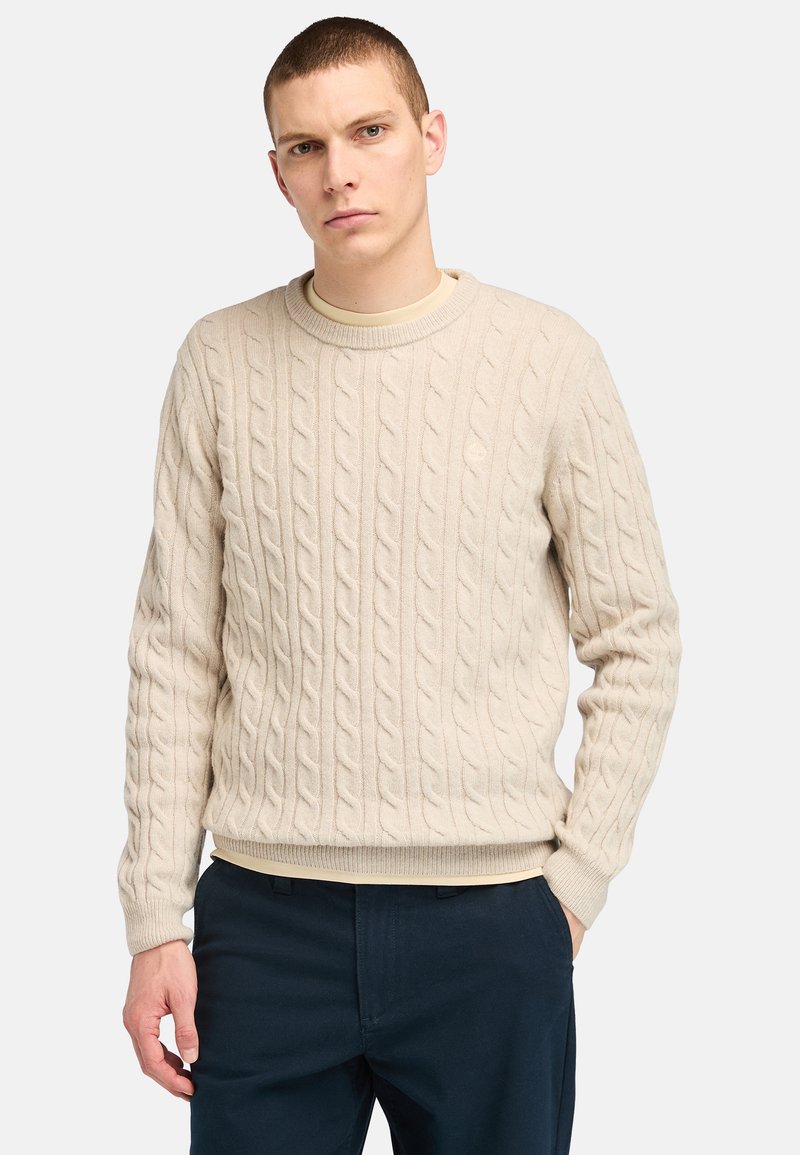 Beige Kabelstrickpullover mit rundem Ausschnitt, gerippten Manschetten und Saum. Über einem hellen Unterhemd getragen, kombiniert mit dunklen Hosen.