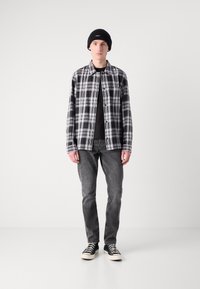 Czarna i biała koszula w kratę na czarnym t-shircie, w połączeniu z szarymi dżinsami i czarnymi trampkami. Model nosi czapkę typu beanie z czarnej wełny.