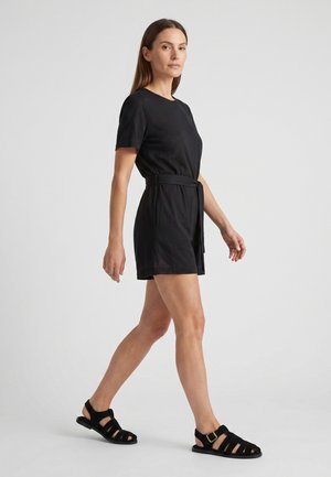 Kvinde går i en sort kortærmet buksedragt med bælte og sorte sandaler med remme mod en ensfarvet lys baggrund.