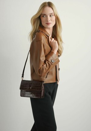 Blondynka w brązowej skórzanej kurtce i czarnych spodniach, trzymająca ciemnobrązową torebkę na ramię o fakturze krokodyla z złotymi okuciami.