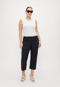 White fitted tank top, black cropped pants, and white strappy sandals. Model wears oversized black sunglasses. Smooth textures, minimalist design.