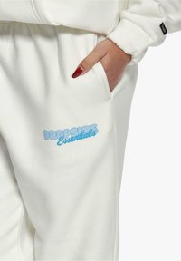 Hvit hettegenser med en forsiktig lomme og en blå "DROPSIZE Essentials" tekstlogo trykt på siden. Laget av myk, strukturert stoff.