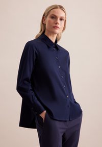 Granatowa koszula z długim rękawem, z kołnierzykiem, guziczkami z przodu i rozcięciami po bokach. Wykonana z gładkiego materiału, o luźnym kroju.