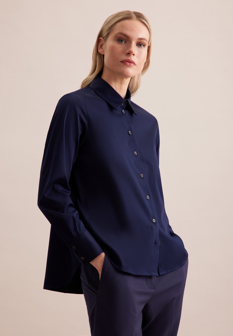 Granatowa koszula z długim rękawem, z kołnierzykiem, guziczkami z przodu i rozcięciami po bokach. Wykonana z gładkiego materiału, o luźnym kroju.