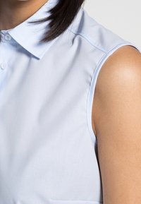 Chemise bleu clair sans manches avec un col pointu, en tissu lisse et avec une légère brillance. Elle comporte une petite poche à l'avant.