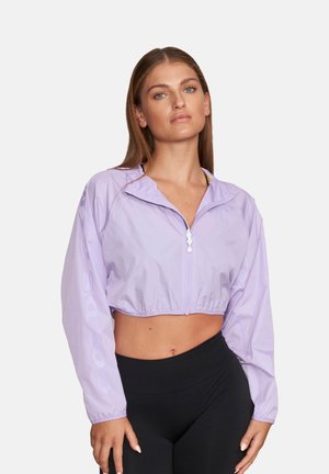 Młoda kobieta z długimi włosami, ubrana w jasnofioletową krótką kurtkę przeciwwiatrową i wysokowysoko taliowane czarne legginsy na jednolitym tle.