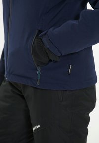 Marinblå jacka med dragkedjefickor och en justerbar snodd vid midjan, i kombination med svarta isolerade byxor med vit logotyp.