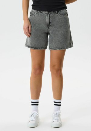 Persoană purtând pantaloni scurți din denim gri, adidași albi și șosete albe cu două dungi negre, stând în fața unui fundal simplu.