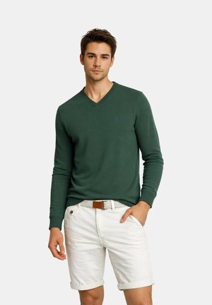 Junger Mann trägt dunkelgrünen V-Ausschnitt-Pullover und weiße hochgekrempelte Shorts mit gewebtem Gürtel, steht mit einer Hand in der Tasche.