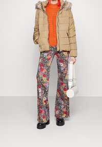 Kvinde iført beige pelshettejakke, orange strikket sweater, farverigt blomsterbukser, sorte platform sko, holder en hvid cylindrisk håndtaske.