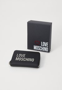 Carteira preta com fecho zip, apresentando o texto "LOVE MOSCHINO" em prata, embalada numa caixa cinza escura com marcação correspondente e detalhe de coração vermelho.