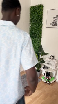 Una persona in camicia azzurro chiaro si trova di fronte a un angolo con una pianta verde, una macchina da cucire su un piccolo tavolo bianco, attrezzatura fotografica e opere d'arte appese al muro con testi motivazionali.