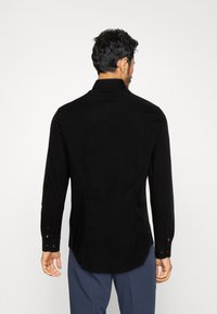 Camisa negra de manga larga con cuello estándar, textura suave, diseño ajustado y puños abotonados; parte trasera lisa sin patrones ni acentos.