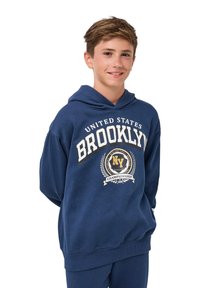 Nastolatek uśmiechający się, ubrany w granatowy polar z napisem "Mistrzostwa Brooklynu NY, Stany Zjednoczone" oraz grafiką laurową z przodu.