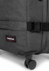 Valigia in tessuto grigio con fibbia nera, patch logo Eastpak e ruote nere, con esterno testurizzato e design aerodinamico.