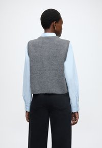 Szara wełniana kamizelka o krótkim kroju, noszona na jasnoniebieskiej koszuli z długim rękawem. Charakteryzuje się fakturowanym wykończeniem i szwami na ramionach.