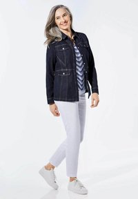 Donkerblauwe denimjas met knopen, twee borstzakken, gedragen over een gestreept shirt en gecombineerd met witte broek en sneakers.