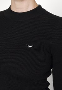 Nahaufnahme einer Person, die einen schwarzen, gerippten Pullover mit einem kleinen Levi's-Logo-Patch auf der linken Brust trägt.